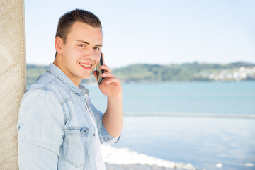 Portrait of happy young man talking on mobile phone outdoors. Smiling guy standing leaning wall and using cellphone at seaside. Mobile communication concept