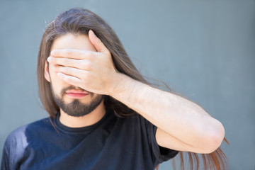 Disappointed young man making face palm gesture. Serious guy with long beautiful hair covering eyes with hand. I do not look concept