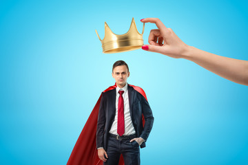 Female hand holding golden crown above young businessman with red cloak on blue background