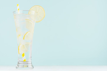 Homemade diet transparent lemonade with lemon, ice cubes, soda, yellow striped straw in misted glass on white wood table, pastel green color background.