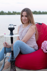 Fototapeta premium Young caucasian beautiful happy woman sitting on a red ottoman and smoking hookah on the city beach in the summer. Chill and relax concept