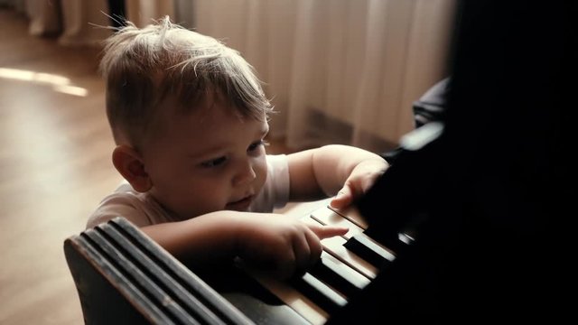 Little cute baby boy with smile on his face knocking on piano keys in slow motion. Child trying to play piano at home. Curious little guy.