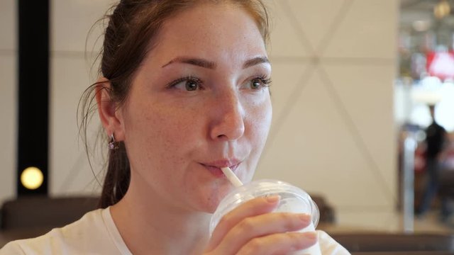Woman is having a luch in fast food restaurant. She is drinking milkshake from straw in cafe. Portrait of young brunette girl.