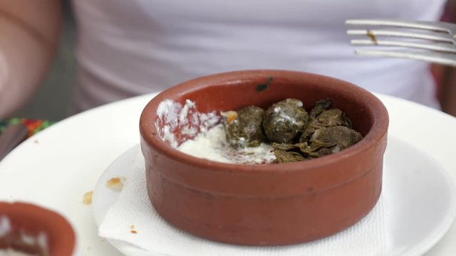 National traditional cuisine food in the restaurant. Woman is eating dolma with sour cream served in clay bowl, dish closeup. Girl is eating with fork putting the sour cream using spoon.