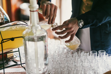 Waiter pouring vodka in glass on table at wedding reception. Guests hands holding lemonade or cocktail from big jar at alcohol bar at wedding or christmas feast. Luxury catering - obrazy, fototapety, plakaty