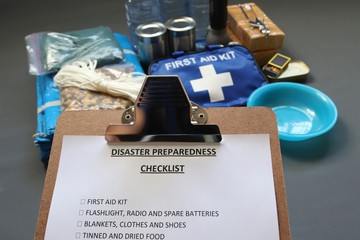 Disaster preparedness checklist on a clipboard with disaster relief items in the background.Such items would include a first aid kit,flashlight,tinned food,water,batteries and shelter.