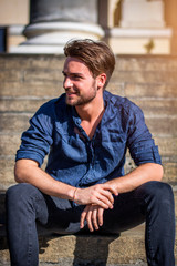 Fototapeta premium One handsome young man in urban setting in summer day, wearing blue shirt, sitting on stone steps