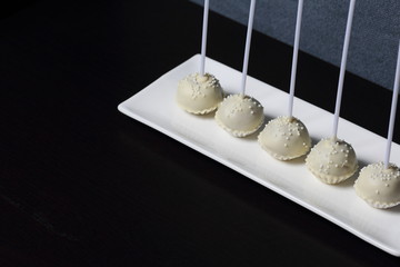 Cake pops in white chocolate glaze. Stand vertically on a stand. Decorated with white decorative sprinkles. On a gray and dark background.