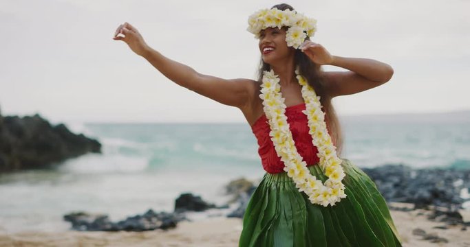 Beautiful smiling woman dancing Hawaiian Hula 'Auana on the beach in slow motion wearing a ti leaf skirt, plumeria lei, and haku