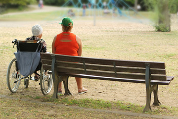woman in an orange jacket and an old woman are disabled in a wheelchair, sit together in a park in summer