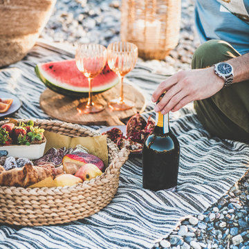 Summer beach picnic at sunset. Young couple sitting on blanket having weekend picnic outdoor at seaside with fresh seasonal fruit, tray of tasty appetizers and bottle of sparkling wine, square crop