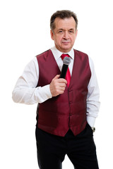 Portrait of successful senior man 55-60 years old. Male stand in beautiful vest with red tie and white shirt. Grinning male hold microphone and second hand in pocket. Isolated on white background.