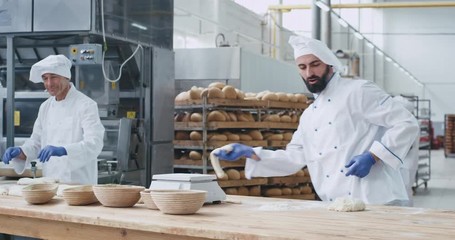 Charismatic very enthusiastic young baker dancing and working in a big bakery industry while forming the dough background workers moving the shelves with bread