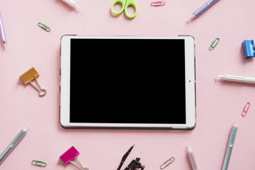 Digital tablet surrounded by various stationeries on pink background