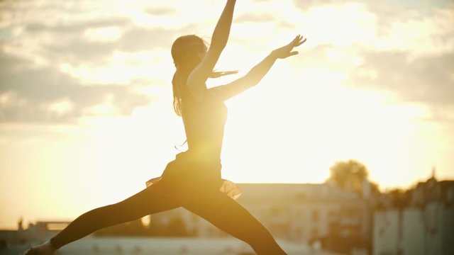 Young woman ballerina in red dress dancing on the roof - runs up and jump performing a split - bright sunset
