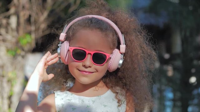 Portrait of a Cute Child, a Wonderful Little Beautiful Girl in a White Dress With Pink Glasses and Pink Headphones, Looking at the Camera, Listening to Music. Concept Happy Childhood.