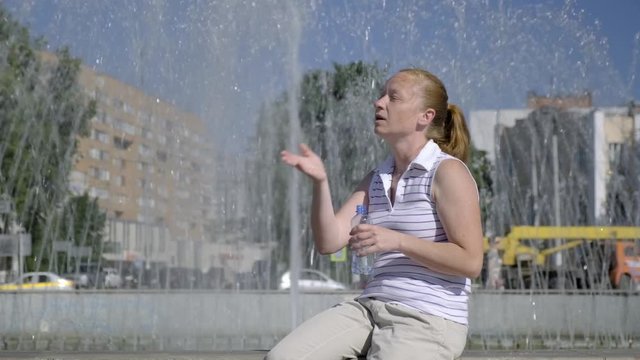 girl suffers from heat at city fountain. Exhausted Woman waving her arms and drinking water from bottle.