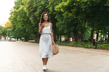 Portrait of happy woman smiling and holding cellphone while walking through green boulevard