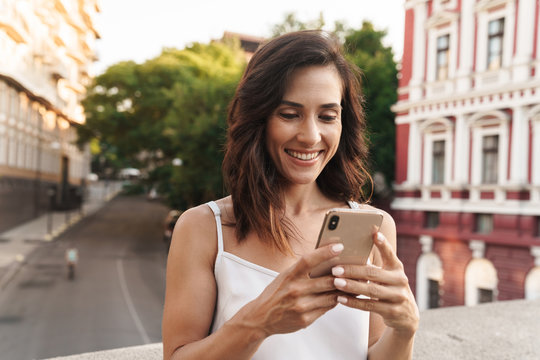 Portrait of cute woman typing on smartphone while standing on bridge with city street background