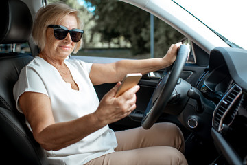Senior business woman using smart phone while driving a car, view from the inside of a vehicle. Concept of an active people during retirement age