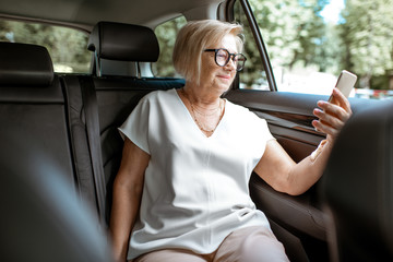 Senior business woman talking with phone while sitting on the back seat in the modern car. Concept of a business life on retirement