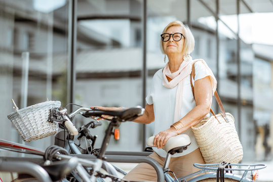 Elegant senior woman parking a bicycle near the modern building outdoors. Concept of an active lifestyle on retirement age