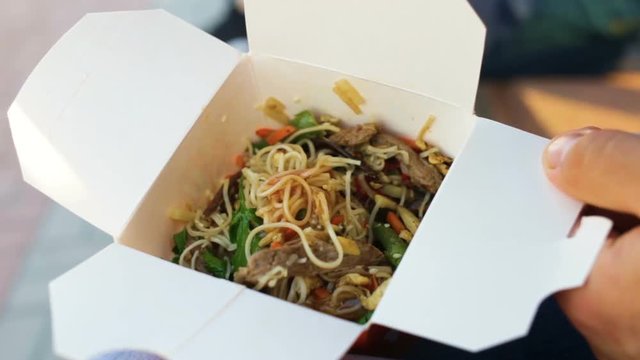 Delicious egg noodles wok in a cardboard disposable dropbox, etc close up. Real time, outdoor, male hands and disposable plastic fork in the frame, at sunset.