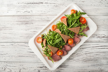 Baked ham with arugula and bread croutons close-up. Snack from jamon, arugula and cherry on a white plate copy space.