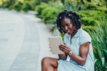 Portrait of an African  smiling young black woman sitting in a park and using a digital tablet  looking at screen