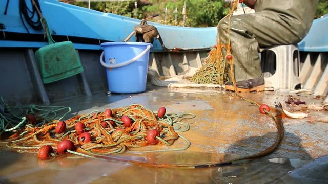 Fisher in rubber trousers and boot siting in his boat and pile up fishing net for angling at open sea. Photo - JPEG video codec