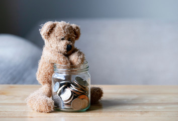 Teddy bear putting hand into the jar of British coins on wooden table , Brown bear sitting next to...