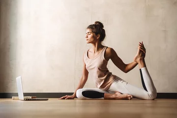  Foto van een jonge geconcentreerde vrouw die yoga-oefeningen doet met een laptop terwijl ze thuis op de vloer zit © Drobot Dean
