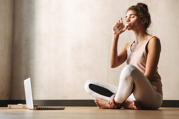Photo of young pretty woman drinking water and sitting on floor while doing yoga exercises at home