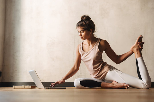 Photo of attractive concentrated woman doing yoga exercises using laptop while sitting on floor at home