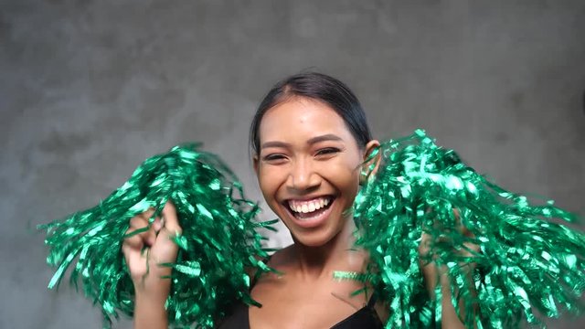 Portrait of beautiful young smiling girl with green cheerleader pom-poms over concrete wall background.
