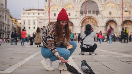 Fototapeta premium Young smiling woman squats in the middle of square and feedings pigeons
