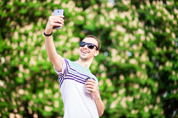 Cutie man in sunglasses with cup of coffee looking at mobile phone outdoors
