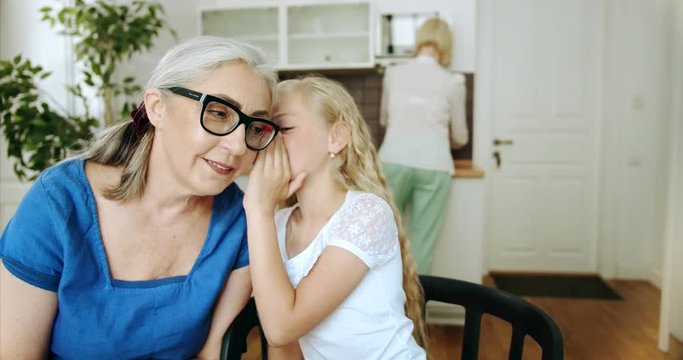 Cute blond girl in white blouse whispering secret to granny's eay, family spending time together indoors