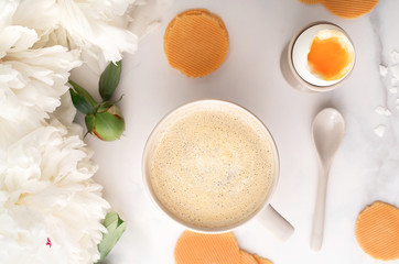Top view of soft-boiled egg with liquide orange yolk in ceramic egg cup, cup of coffee with ceramic spoon, thin crispy corn chips and beautiful white peony. Breakfast concept. Flat lay.