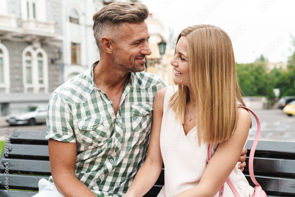 Wall mural Portrait of pleased young couple smiling and hugging together while sitting on bench in city street