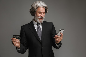 Image of happy adult businessman wearing formal black suit holding credit card and smartphone