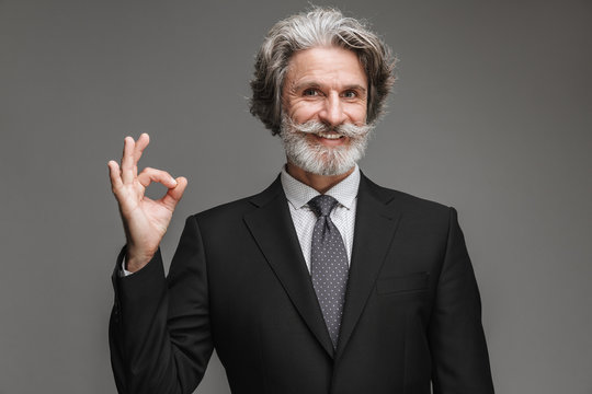 Image of joyful adult businessman wearing formal black suit smiling at camera and showing ok sign