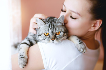Woman at home kissing and hug her lovely fluffy cat. Gray tabby cute kitten with yellow eyes. Pets, friendship, trust, love, and lifestyle concept. Friend of human. Animal lover. Close up.