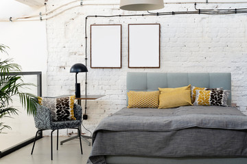 Industrial interior of bedroom in scandinavian style with grey bed, pillows and white bricky wall with blank frames with mock up.
