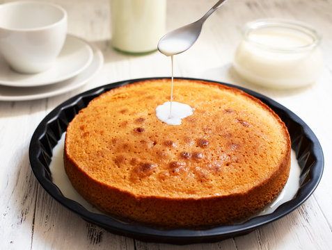 Traditional dessert Three Milk, Tres Leches or Trilece. Served on a white wooden table with milk in a bottle and a cup. Milk pours from a spoon onto a biscuit. Biscuit soaked with milk. Closeup