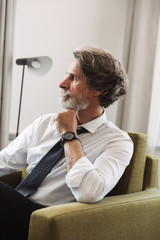 Image of handsome mature man wearing suit and tie sitting on armchair in bright apartment