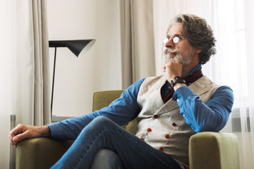 Image of confident adult professor man wearing stylish suit and eyeglasses while sitting on armchair in hotel apartment