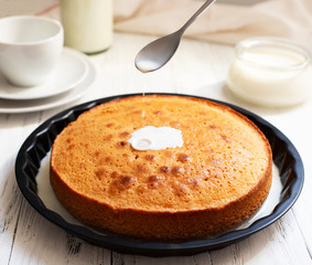 Traditional dessert Three Milk, Tres Leches or Trilece. Served on a white wooden table with milk in a bottle and a cup. Milk pours from a spoon onto a biscuit. Biscuit soaked with milk. Closeup