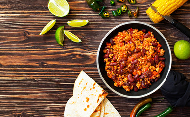 Traditional Mexican chili con carne served on a rustic wooden background in a pan with corn, mexican tortilla bread, lime and jalapeño. Top view. Space for text