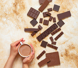 Female hands with a cup of cocoa on a light marble background. Assortment of different types of chocolate. Top view, flat lay. Space for text